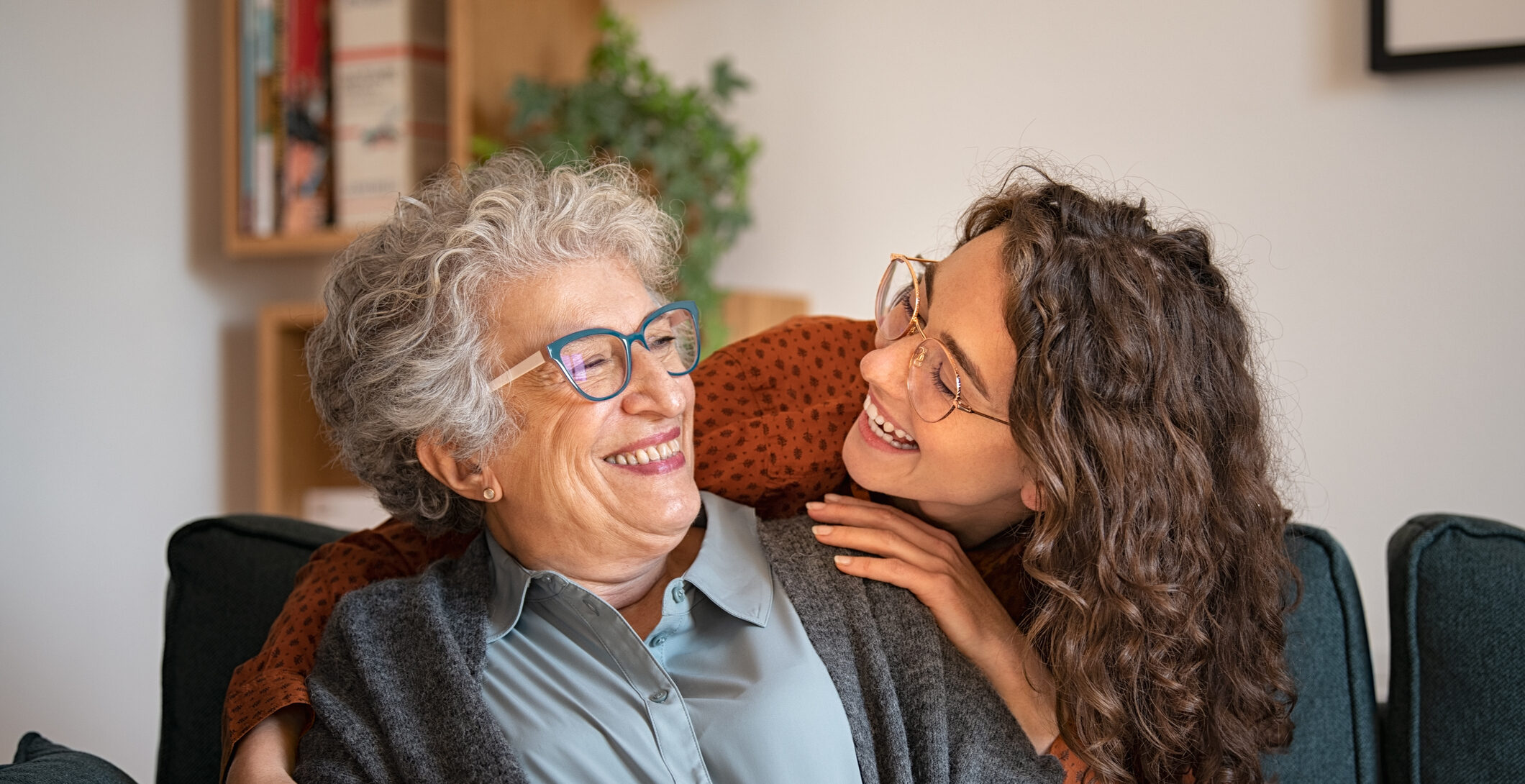 Old grandmother and adult granddaughter hugging at home and looking at each other. Happy senior mother and young daughter embracing with love on sofa. Happy young woman hugging from behind grandma with love.