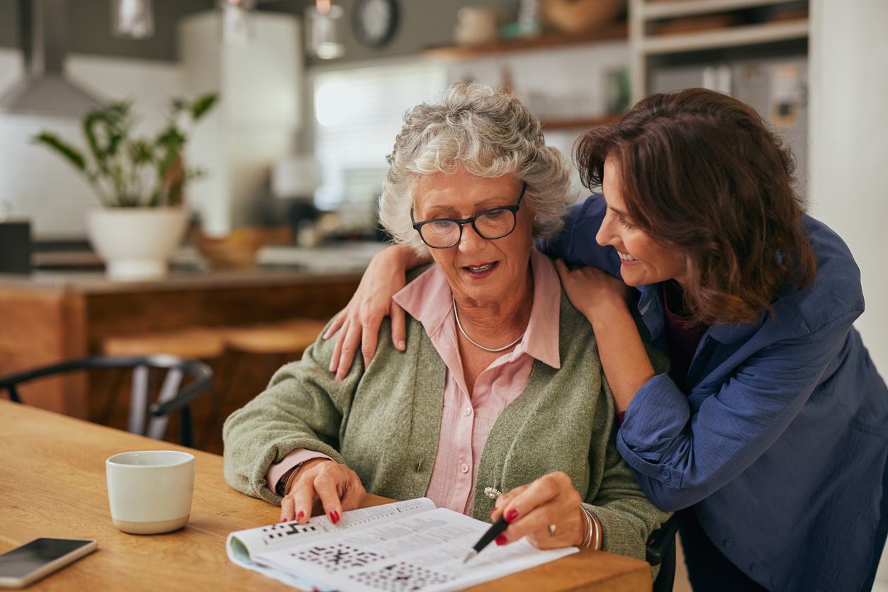 Senior woman working on a crossword puzzle in newspaper with adult daughter helping. Elderly mother and daughter bonding while solving a crossword puzzle together at home with copy space. Smiling mother and young woman keeping mind trained through memory exercises.