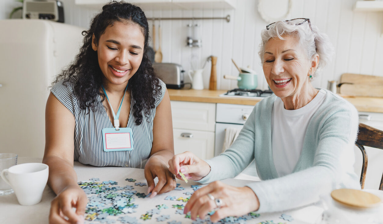 Indoor portrait of young black female volunteer and Caucasian old lady collecting puzzle together at kitchen table, having fun, talking and drinking tea. Social support for lonely elderly people