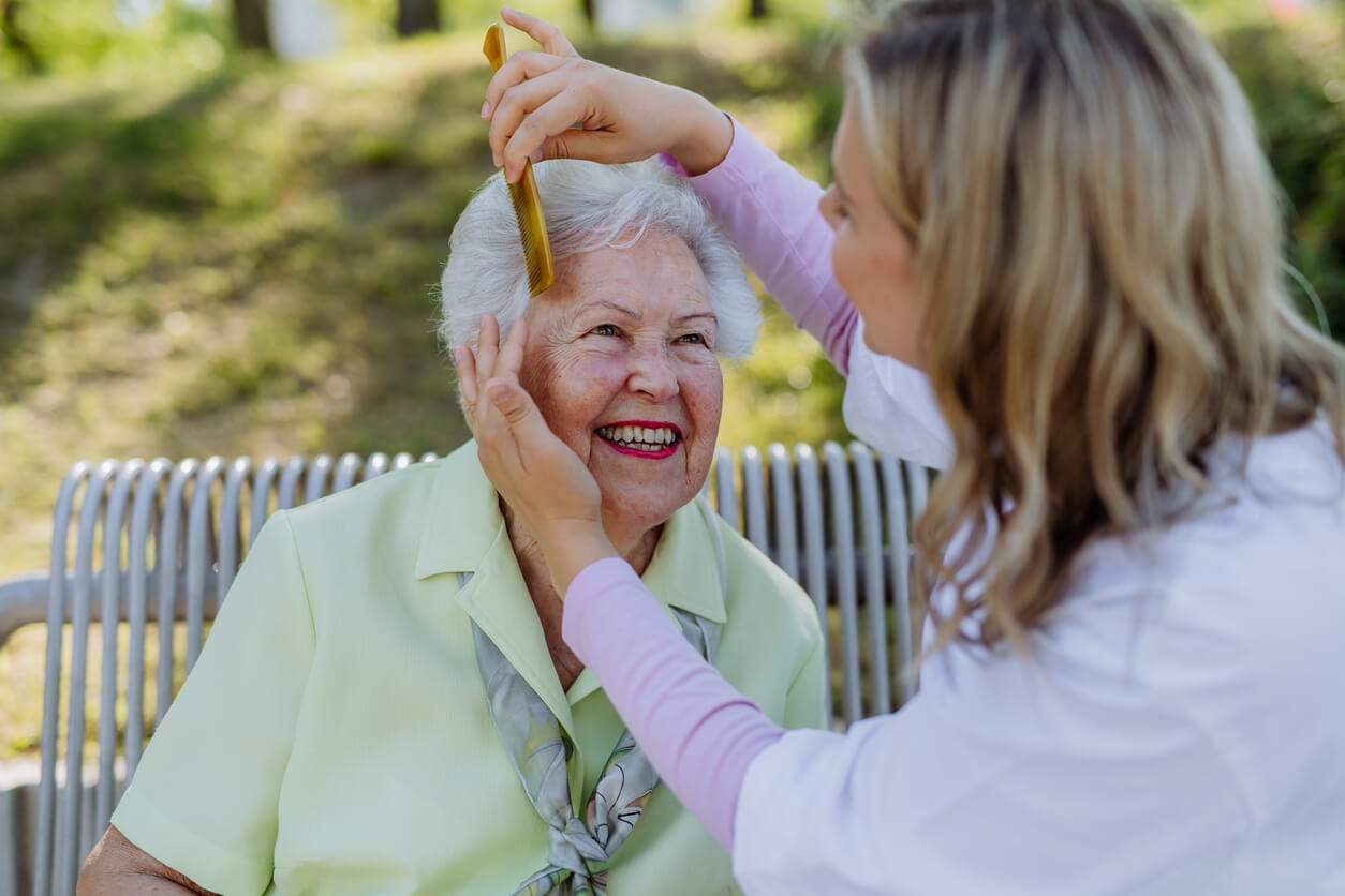 Caregiver helping senior woman to comb hair and make hairstyle when sitting on bench in park in summer.