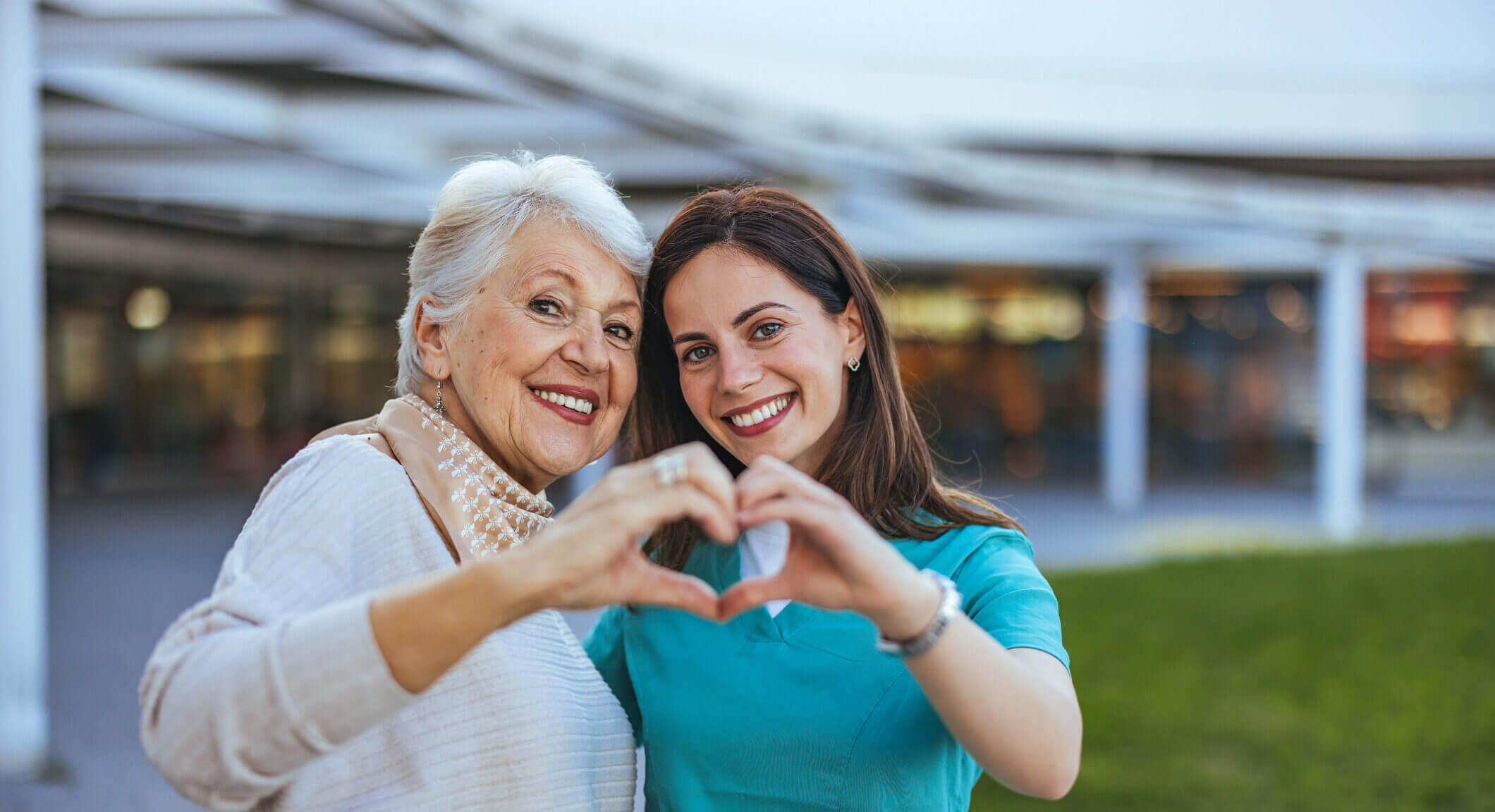 Elderly woman and her caregiver happily form a heart shape with their hands.